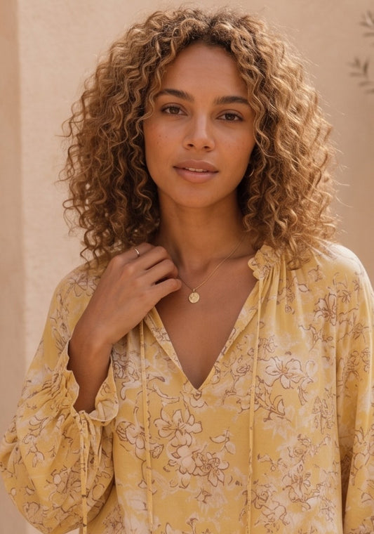 blusa de estilo bohemio de color amarillo vibrante con un delicado estampado floral en tonos blancos y marrones. La blusa es de tela fluida y ligera, perfecta para los días calurosos, y presenta detalles como un cuello redondo con un pequeño escote en V que se cierra con un cordón y borlas finas. Las mangas son largas y abullonadas, lo que le da un aire romántico y femenino