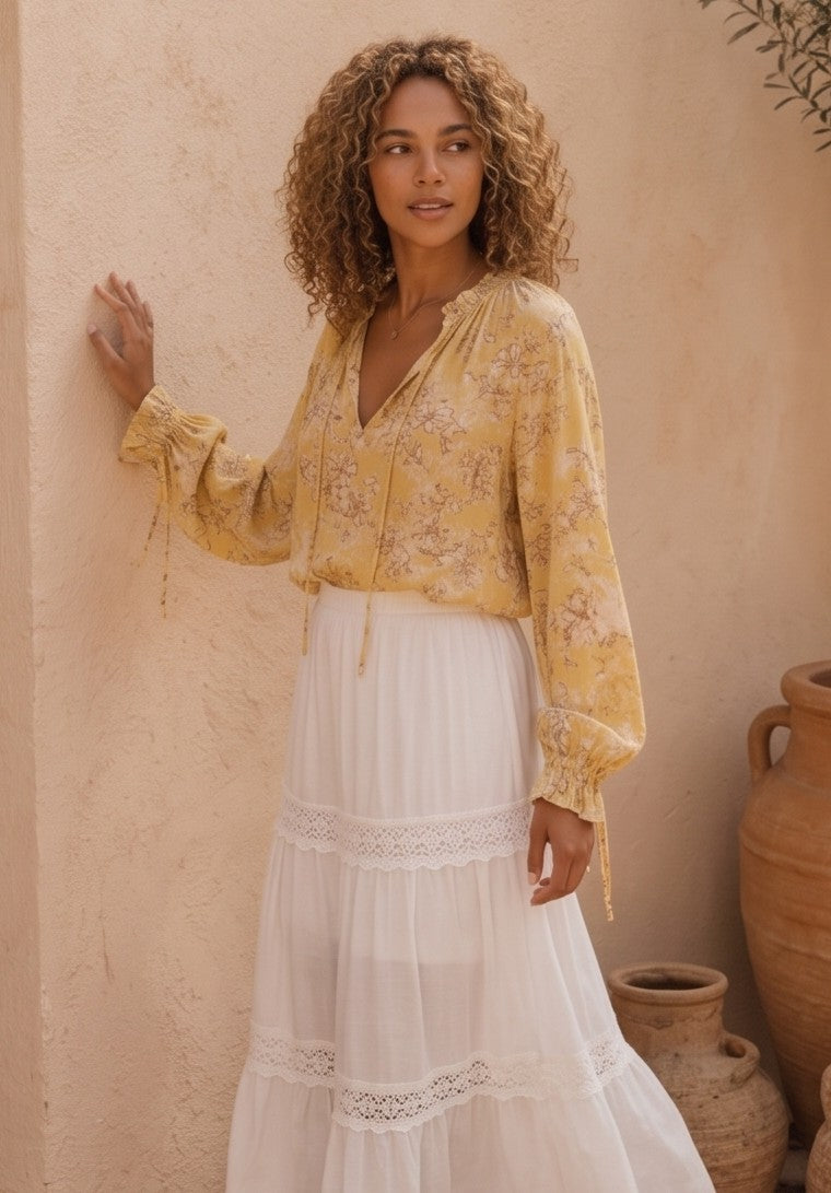 blusa de estilo bohemio de color amarillo vibrante con un delicado estampado floral en tonos blancos y marrones. La blusa es de tela fluida y ligera, perfecta para los días calurosos, y presenta detalles como un cuello redondo con un pequeño escote en V que se cierra con un cordón y borlas finas. Las mangas son largas y abullonadas, lo que le da un aire romántico y femenino