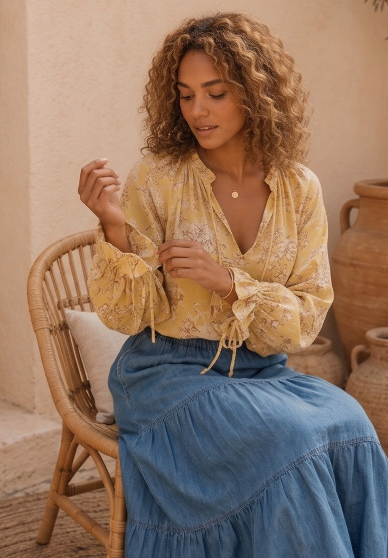 blusa de estilo bohemio de color amarillo vibrante con un delicado estampado floral en tonos blancos y marrones. La blusa es de tela fluida y ligera, perfecta para los días calurosos, y presenta detalles como un cuello redondo con un pequeño escote en V que se cierra con un cordón y borlas finas. Las mangas son largas y abullonadas, lo que le da un aire romántico y femenino