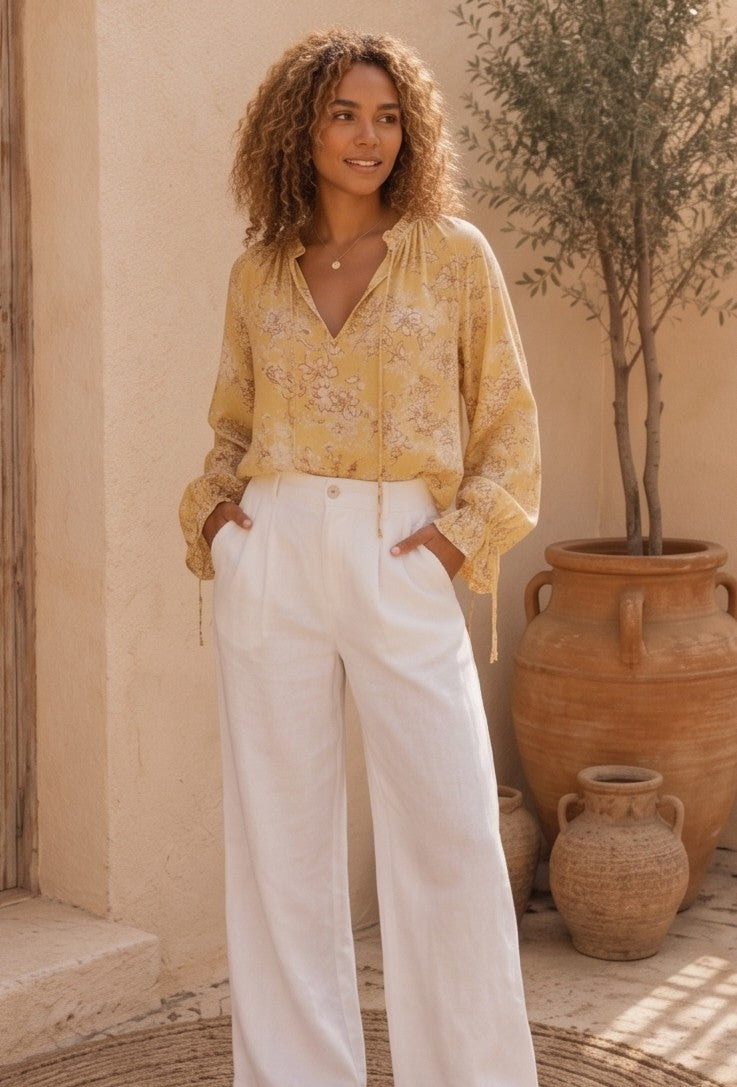 blusa de estilo bohemio de color amarillo vibrante con un delicado estampado floral en tonos blancos y marrones. La blusa es de tela fluida y ligera, perfecta para los días calurosos, y presenta detalles como un cuello redondo con un pequeño escote en V que se cierra con un cordón y borlas finas. Las mangas son largas y abullonadas, lo que le da un aire romántico y femenino