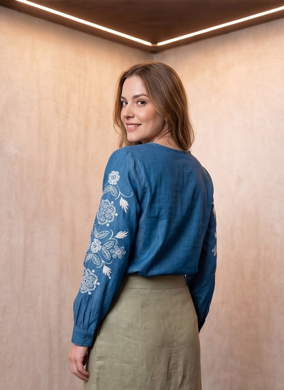 Blusa vaquera azul denim de manga larga con bordados florales blancos en las mangas y cuello redondo abotonado.