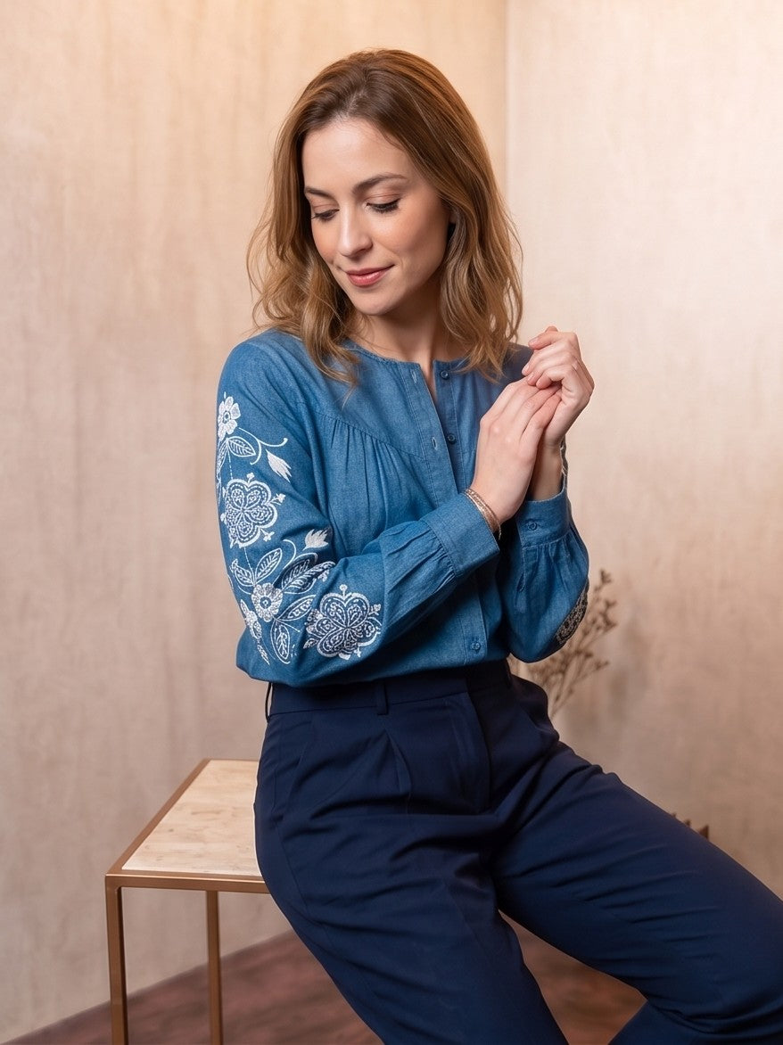 Blusa vaquera azul denim de manga larga con bordados florales blancos en las mangas y cuello redondo abotonado.