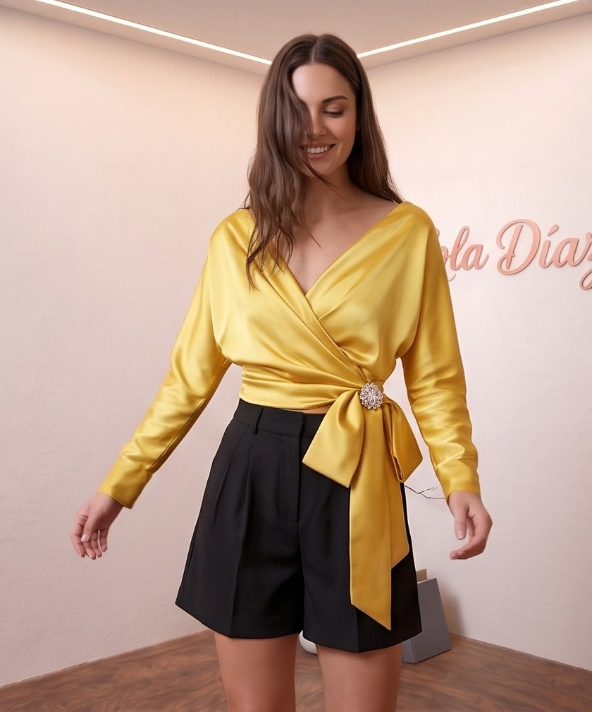 Blusa de satén de seda con diseño cruzado, escote en V y lazada lateral con detalle de broche. Disponible en varios colores: amarillo vibrante, azul cielo, blanco roto y champán. Manga larga y corte elegante y sofisticado.