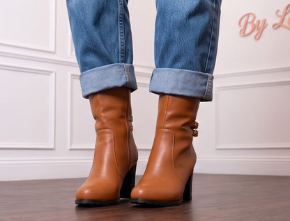Botas de media caña para mujer en polipiel PU con tacón ancho alto y detalles metálicos dorados en la base del tacón. Diseño de puntera redonda con doble tira lateral decorativa, hebillas doradas redondas y cierre de cremallera interior. Disponibles en color negro, blanco y camel