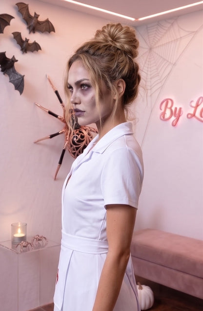 Disfraz de enfermera para mujer en color blanco liso, confeccionado en poliéster. Incluye vestido corto con cuello camisero y escote en pico, cofia o tocado a juego para la cabeza con detalle de cruz roja y delantal blanco ajustable con cruz médica en el pecho