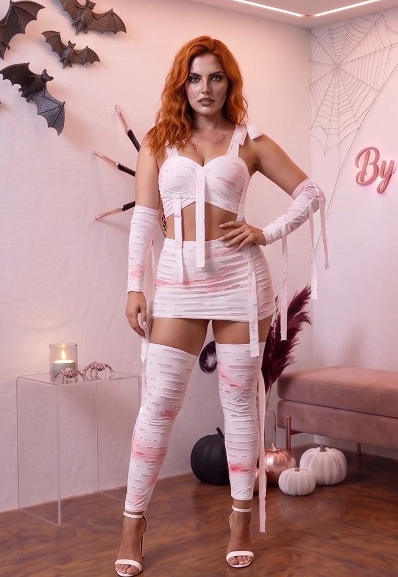 Disfraz de momia para mujer de cuatro piezas en tela elástica blanca con efecto de tiras desgarradas, roturas y manchas de sangre artificial rosa y roja. El conjunto incluye un crop top cruzado con tirantes ajustables, falda corta a juego con cintas colgantes, mangas largas y calentadores cubrepiés a juego sobre fondo de estudio de Halloween.
