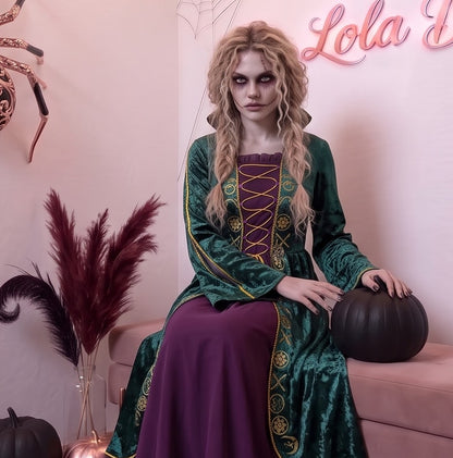 Disfraz de bruja medieval inspirado en Winifred Sanderson con vestido largo en dos piezas simuladas: túnica exterior de terciopelo verde esmeralda con cuello alzado, mangas acampanadas con ribetes dorados y bordados rúnicos en el pecho, sobre falda interior fluida en tono morado púrpura con corsé ajustable de cordones amarillos.
