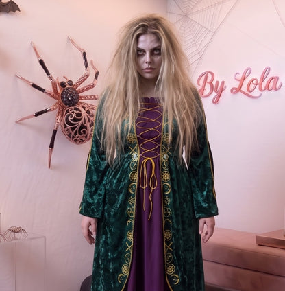 Disfraz de bruja medieval inspirado en Winifred Sanderson con vestido largo en dos piezas simuladas: túnica exterior de terciopelo verde esmeralda con cuello alzado, mangas acampanadas con ribetes dorados y bordados rúnicos en el pecho, sobre falda interior fluida en tono morado púrpura con corsé ajustable de cordones amarillos.