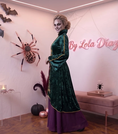 Disfraz de bruja medieval inspirado en Winifred Sanderson con vestido largo en dos piezas simuladas: túnica exterior de terciopelo verde esmeralda con cuello alzado, mangas acampanadas con ribetes dorados y bordados rúnicos en el pecho, sobre falda interior fluida en tono morado púrpura con corsé ajustable de cordones amarillos.