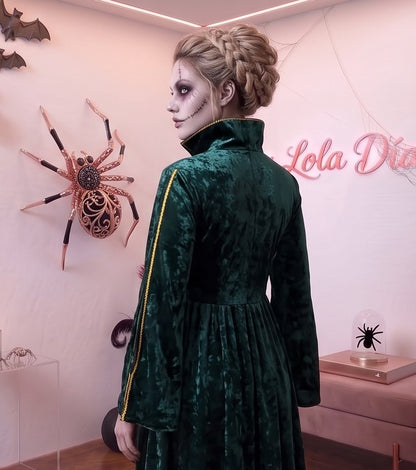 Disfraz de bruja medieval inspirado en Winifred Sanderson con vestido largo en dos piezas simuladas: túnica exterior de terciopelo verde esmeralda con cuello alzado, mangas acampanadas con ribetes dorados y bordados rúnicos en el pecho, sobre falda interior fluida en tono morado púrpura con corsé ajustable de cordones amarillos.