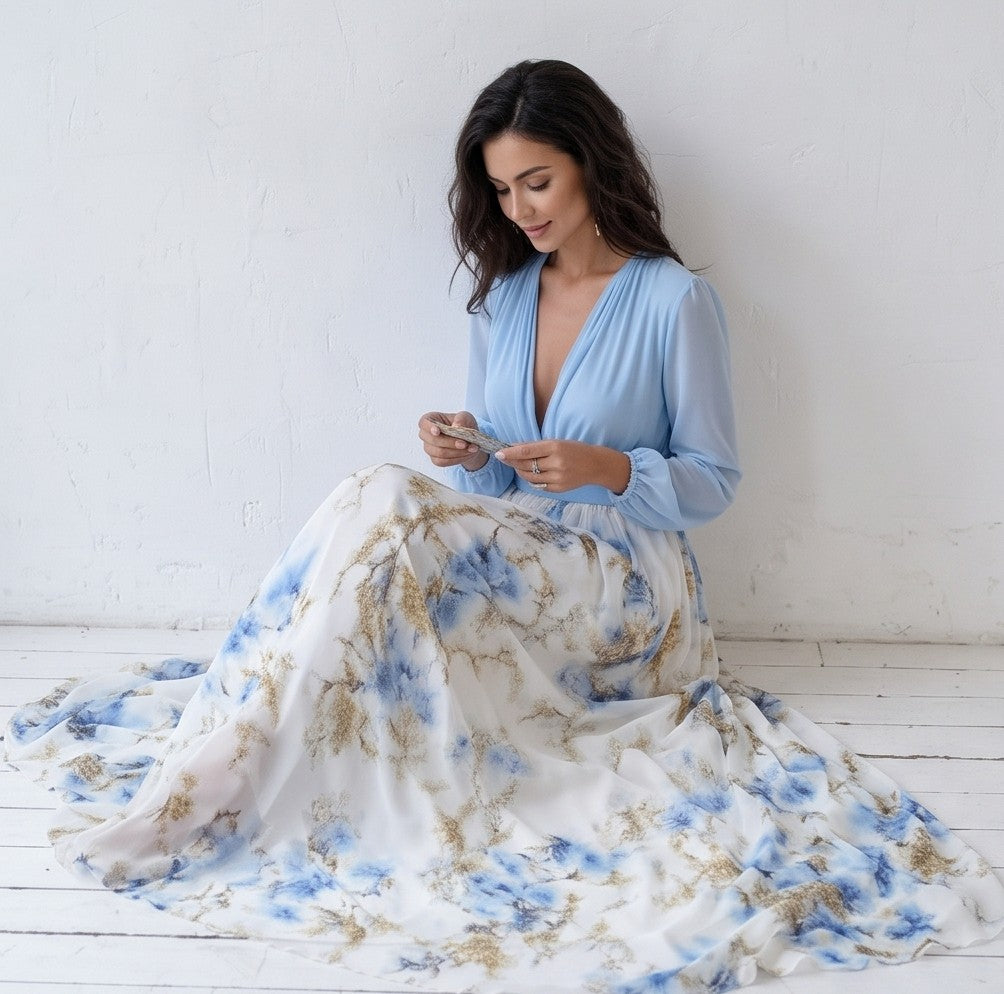 Vestido largo de fiesta para invitada con cuerpo cruzado en gasa azul cielo, escote en V pronunciado, mangas largas abullonadas y falda fluida blanca con estampado floral en tonos azules y dorados.