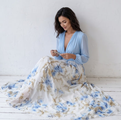 Vestido largo de fiesta para invitada con cuerpo cruzado en gasa azul cielo, escote en V pronunciado, mangas largas abullonadas y falda fluida blanca con estampado floral en tonos azules y dorados.